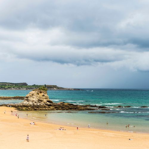 People enjoy Sardinero Beach in Santander on a cloudy day, with waves gently lapping at the shore.