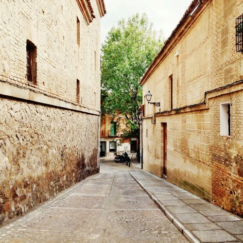 A narrow street in Toledo's Jewish Quarter, lined with historic stone buildings under a clear blue sky.