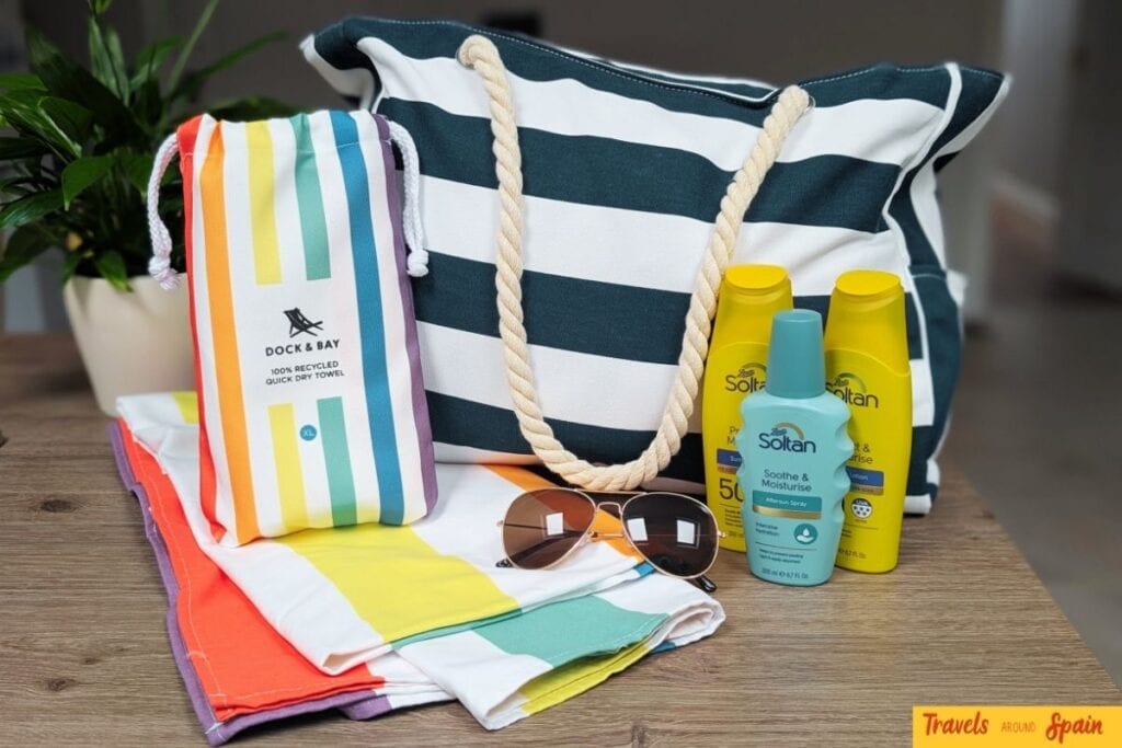 Beach bag with navy and white stripes, colorful striped towels, sunglasses, and Soltan sun protection products arranged on wooden surface for Gran Canaria packing guide