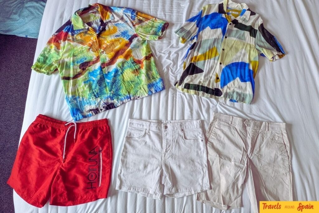 Flat lay of summer clothing showing what to pack for Gran Canaria in June - colorful short-sleeve shirts, swim shorts, and lightweight shorts on white bedding.