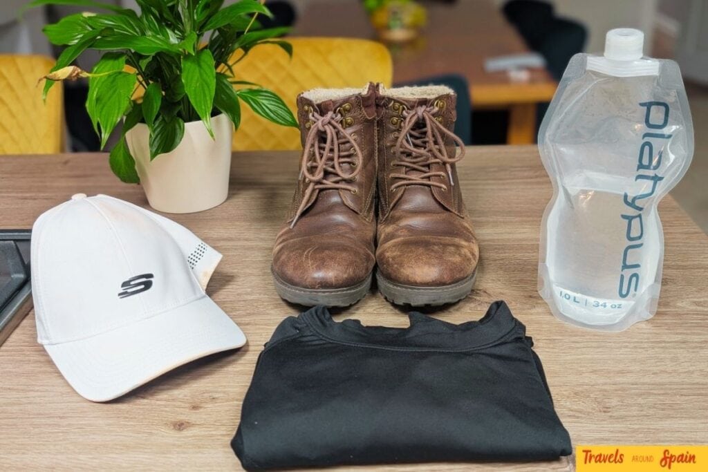 Hiking essentials for what to pack for Gran Canaria in June - brown hiking boots, black long-sleeve shirt, collapsible water bottle and white cap on wooden surface