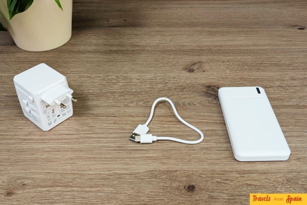 Technology essentials showing what to pack for Gran Canaria in June - universal adapter, charging cable and white power bank on wooden table.