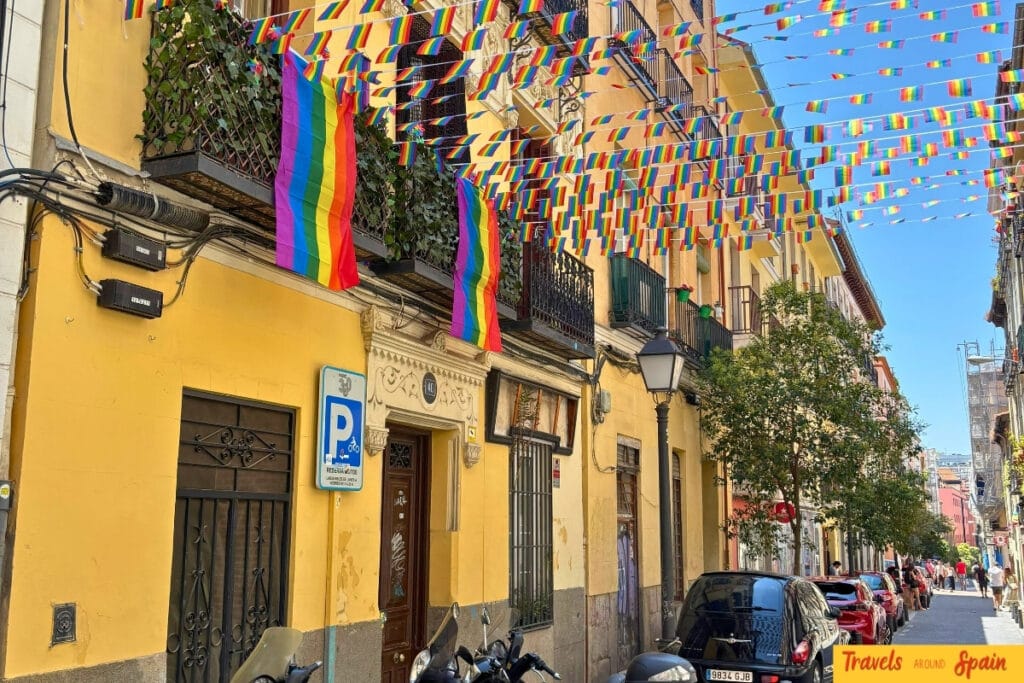 Bright yellow building in Madrid decorated with rainbow pride flags hanging from balconies, with colourful bunting strung overhead across the street under blue skies.