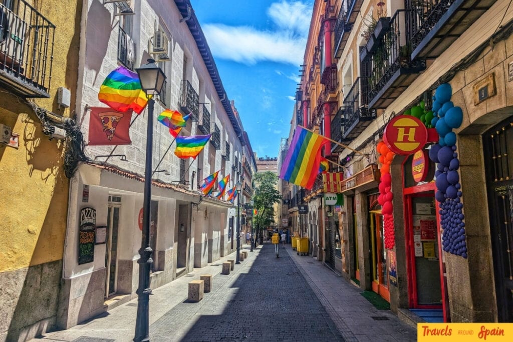 Narrow cobblestone street in Madrid's Chueca district lined with colourful buildings, shops, and numerous rainbow pride flags strung between balconies under a bright blue sky.