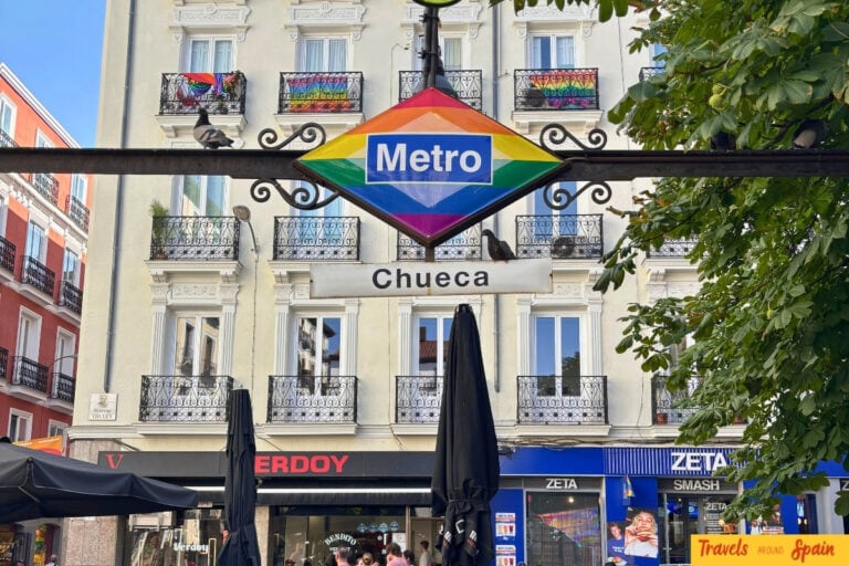 Chueca Metro station sign decorated with rainbow pride colours on its diamond-shaped logo, surrounded by traditional Madrid buildings with wrought iron balconies and street-level shops.