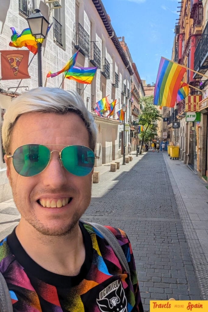 Smiling man in green sunglasses taking a selfie on a cobblestone street in Madrid decorated with rainbow pride flags hanging between historic buildings with wrought iron balconies.
