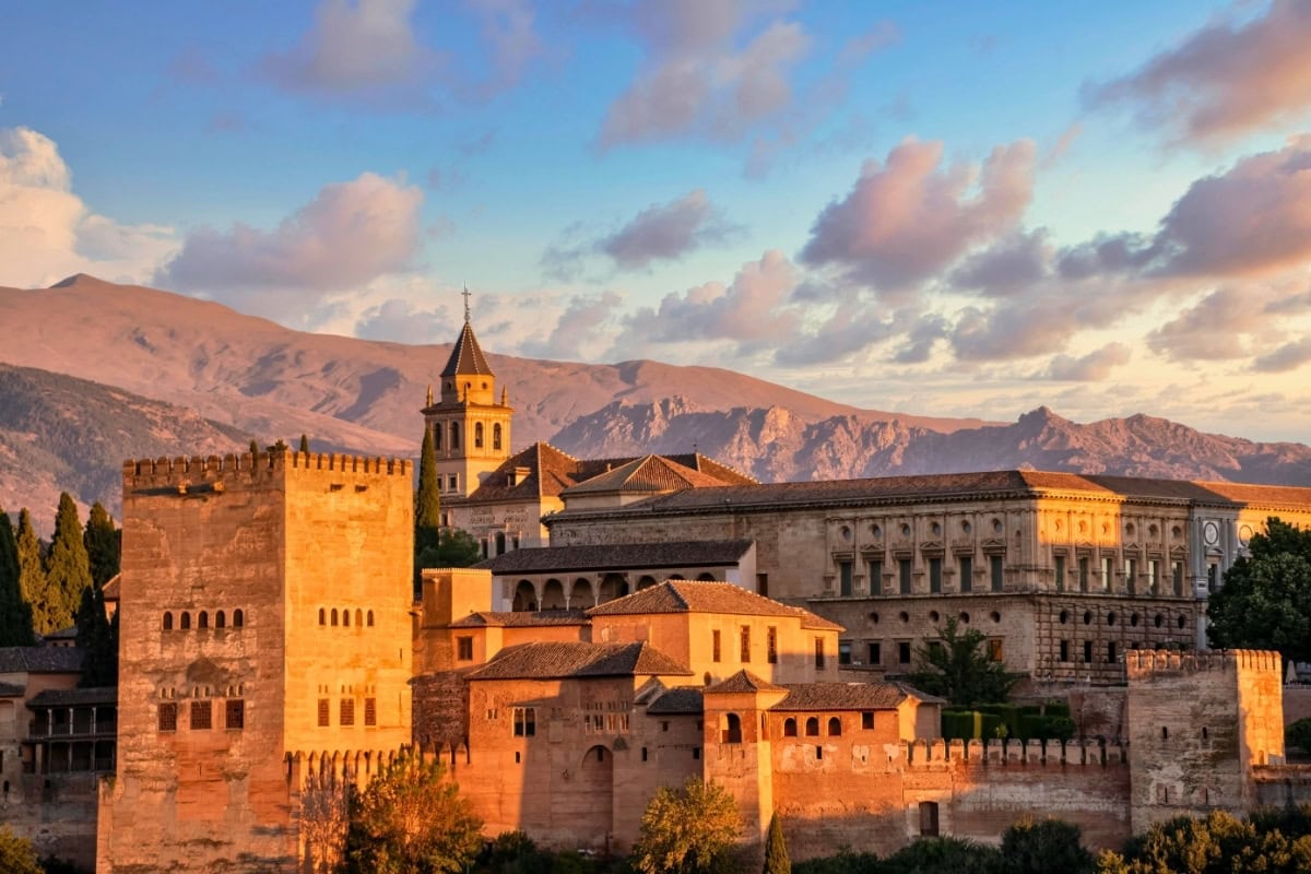 Alhambra fortress illuminated during golden hour sunset with warm orange light on medieval towers and Sierra Nevada mountains beyond.