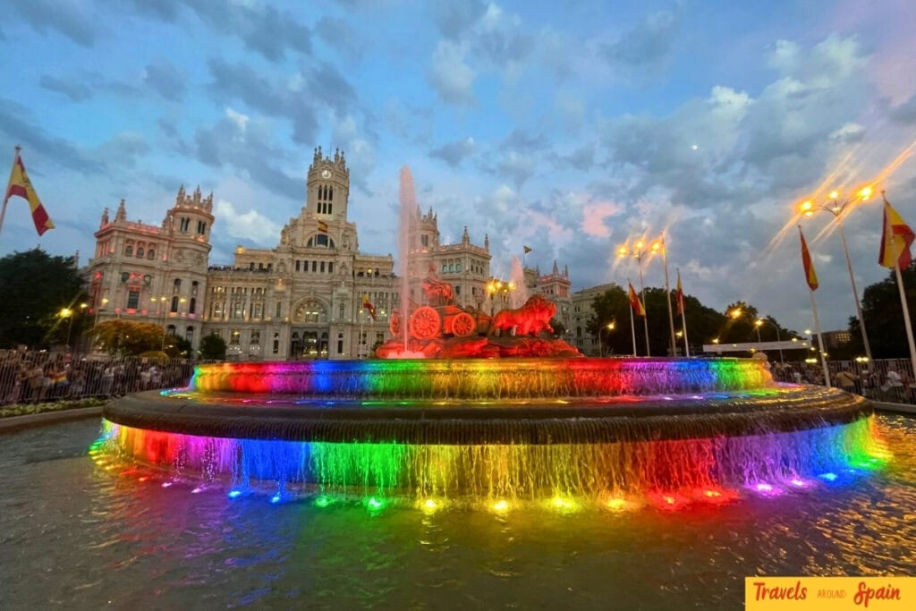 Madrid's iconic Cibeles Fountain illuminated in rainbow pride colours at dusk, with the ornate Cybele Palace and city hall visible in the background under a dramatic cloudy sky.