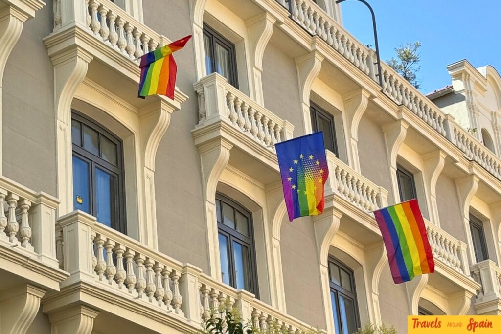 Elegant cream-coloured building with ornate balustrades displaying multiple rainbow pride flags and an EU rainbow flag from its balconies in Madrid.