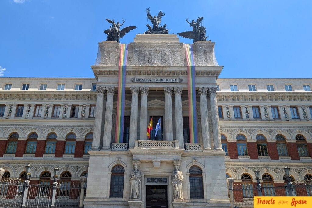 Grand neoclassical government building in Madrid with ornate columns and bronze sculptures, decorated with rainbow pride flags draped across its facade against a blue sky.