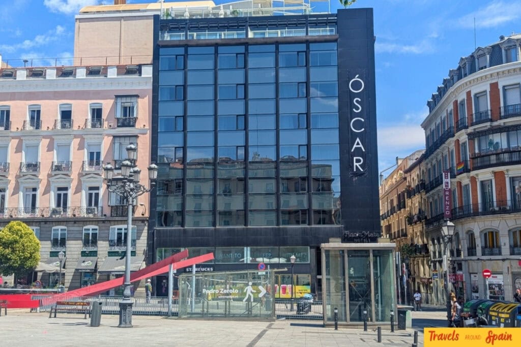 Modern glass building with "ÓSCAR" signage in Madrid's city centre, surrounded by traditional Spanish architecture with ornate balconies and a pedestrian square.