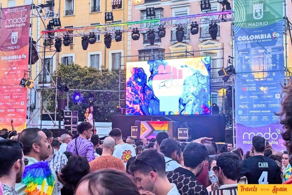 Large crowd at Madrid Pride festival with rainbow stage lighting and historic Madrid buildings