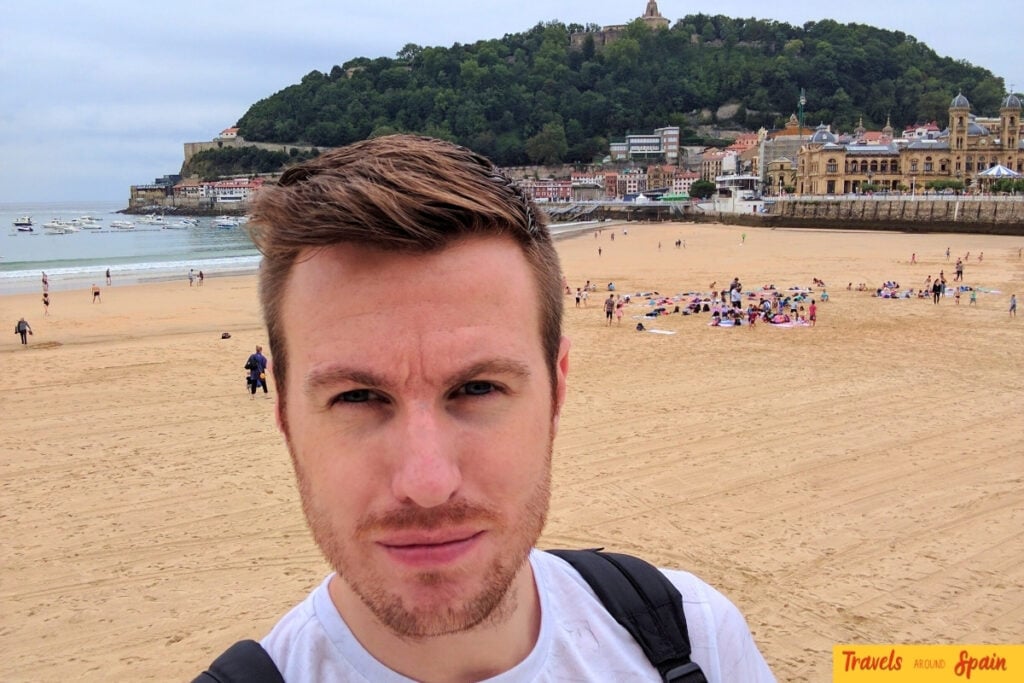 A man with brown hair and facial hair takes a selfie on a sandy beach. Behind him is a picturesque coastal town with historic buildings, a forested hillside, and beachgoers enjoying the shore. The architecture suggests a Spanish Basque coastal location.