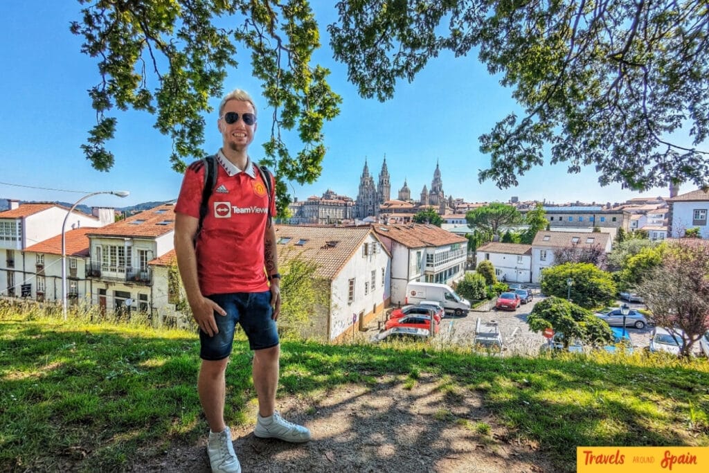 A blonde man wearing sunglasses and a red Manchester United TeamViewer jersey poses on a hillside overlooking a historic Spanish city. Behind him are traditional red-tiled roofs, white buildings, and the distinctive twin spires of Santiago de Compostela Cathedral rising above the cityscape.