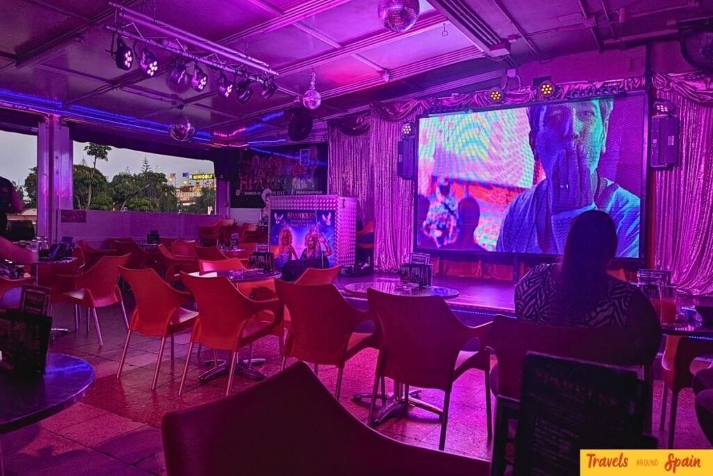Interior of Sparkles Bar Maspalomas showing red plastic chairs, raised stage with LED screen and purple disco lighting.