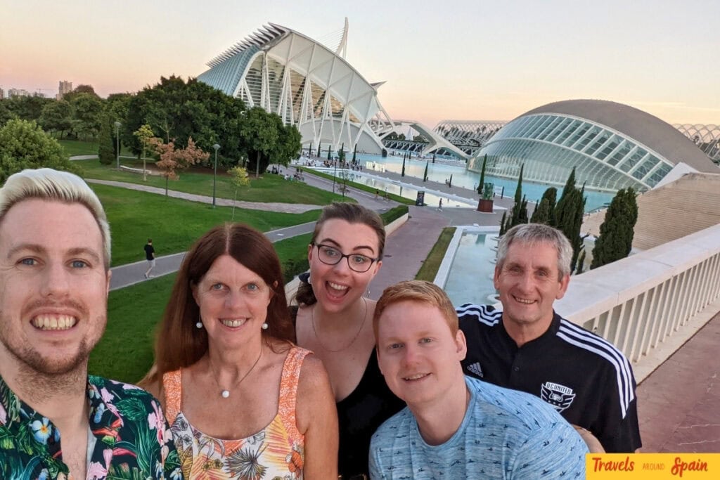 A group of five people take a family selfie at sunset in front of Valencia's futuristic City of Arts and Sciences complex. The modern white architectural structures with their distinctive curved designs are illuminated in the golden hour light, with manicured gardens and walkways visible below.