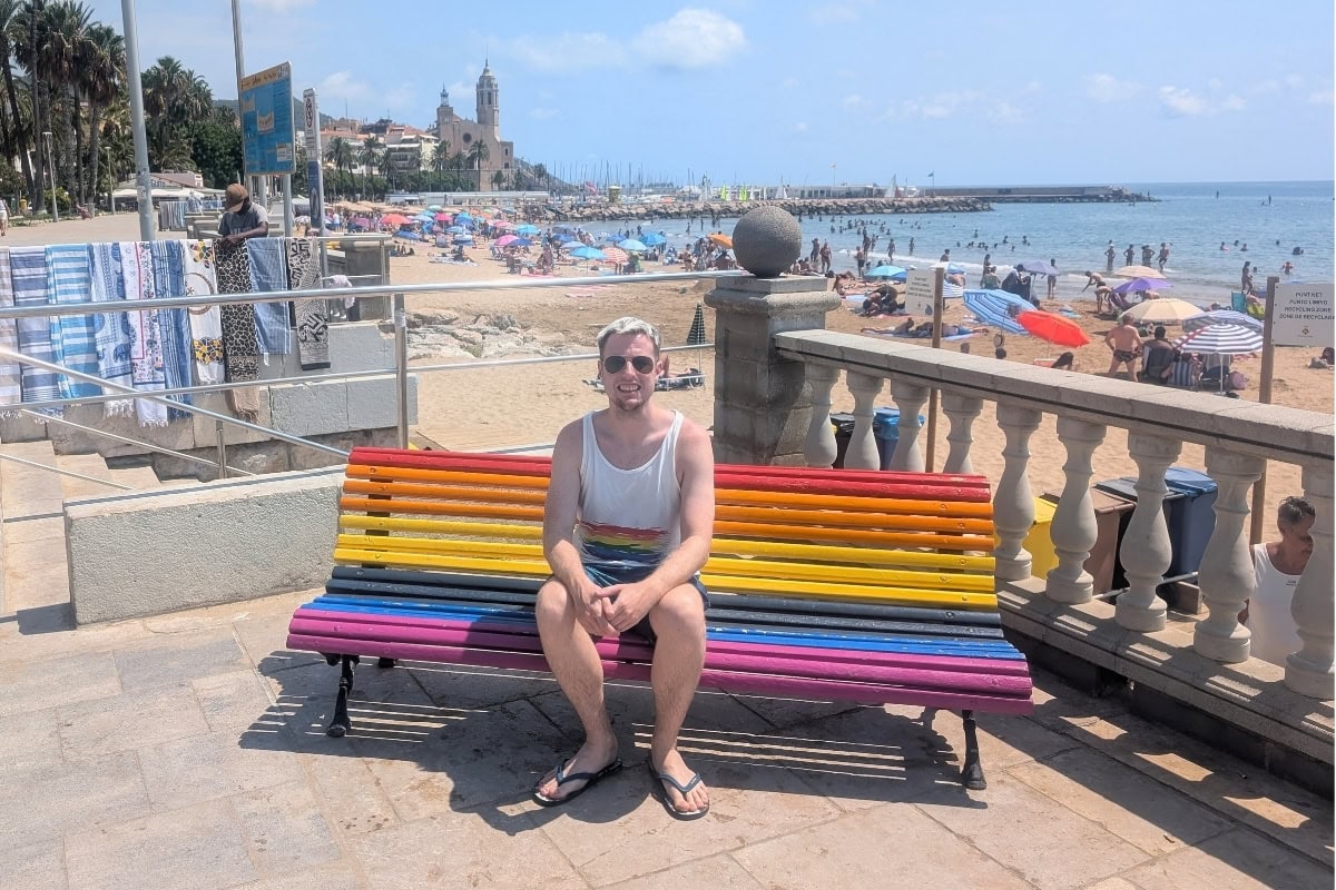 Tourist sitting on rainbow bench at Sitges beach with Sant Bartomeu church and Mediterranean coastline in background