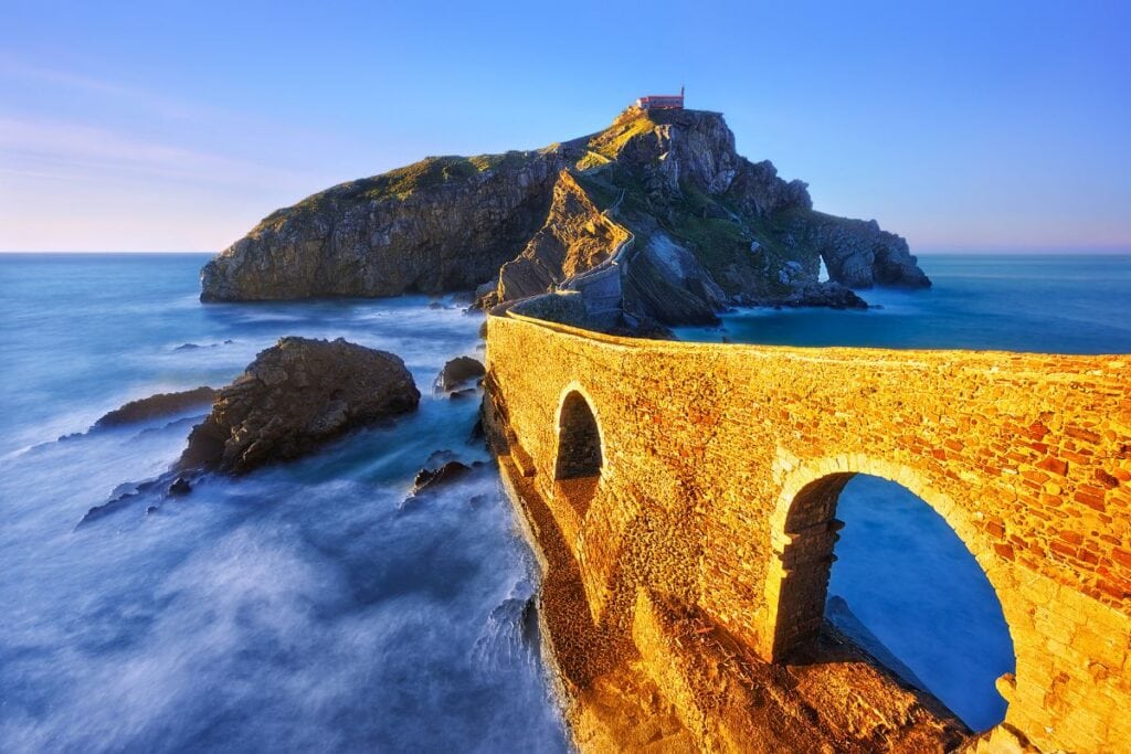 Golden-hour view of San Juan de Gaztelugatxe with its dramatic stone bridge and rocky coastline, capturing the ultimate and most immersive travel experience in Spain.