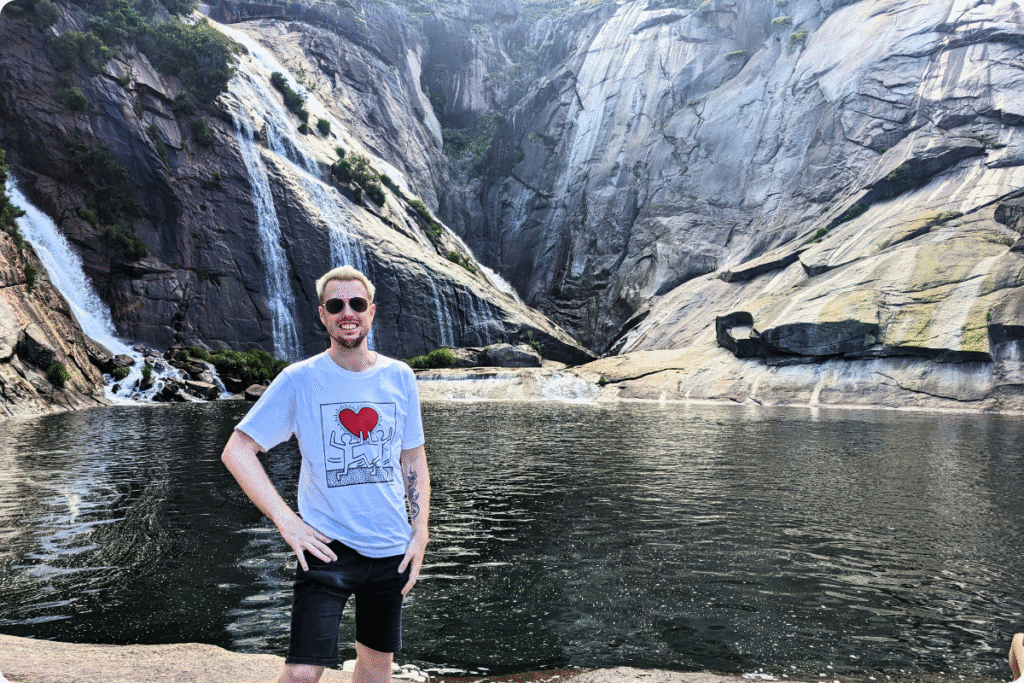 A man poses by the Ézaro Waterfall in Galicia, Spain, where cascading streams flow down rocky cliffs into a tranquil pool.