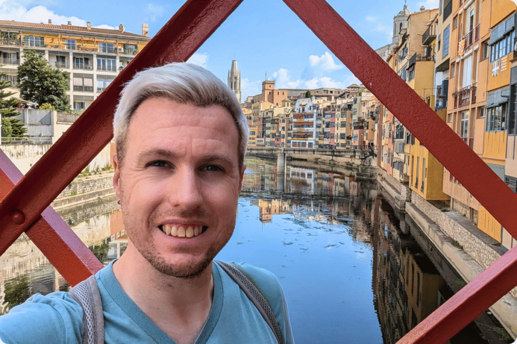 A smiling man takes a selfie on the red Eiffel Bridge with the colourful riverside houses of Girona reflected in the River Onyar.