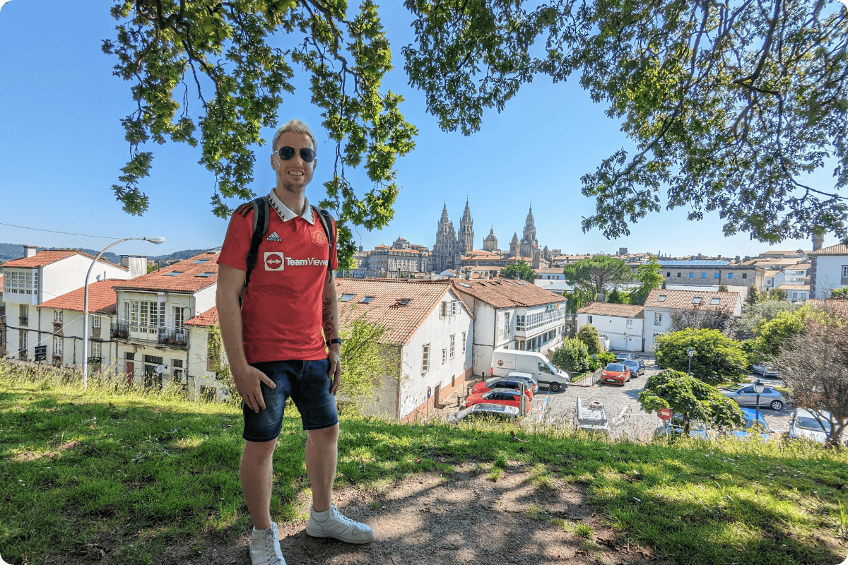 A man wearing a red Manchester United shirt poses on a hill with a panoramic view of Santiago de Compostela Cathedral and the old town in Galicia, Spain.